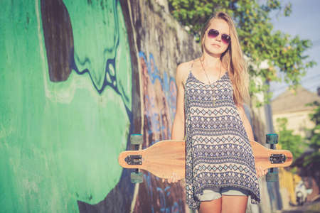 Fashion lifestyle, Beautiful young blonde woman with skateboard standing near the wall with graffiti at the day time.の写真素材