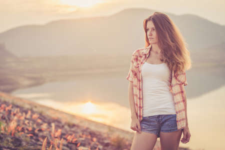Trendy Hipster Girl Relaxing near the lake at the day time. Concept of Modern Youth Lifestyle.  Blowing Long Hair.  Glow Sun, Sunshine. Backlit. Toned in warm colorsの写真素材