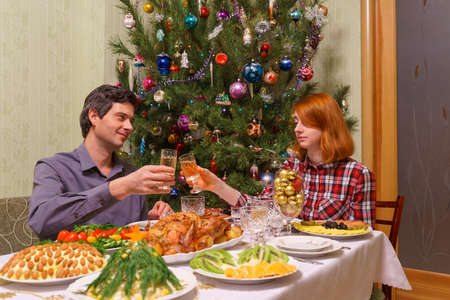 Handsome middle-aged man and beautiful young lady celebrating new year holidays with champagne siitting at festive table near decorated Christmas tree in living room interior. Cozy home conceptの写真素材