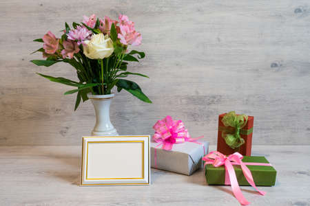 Colorful spring bouquet of rose, chrysanthemum and alstroemeria flowers in a vase with empty photoframe and gift boxes on wooden backgroundの写真素材