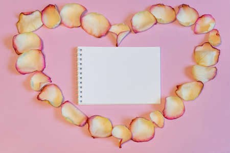 Notebook in frame of dried cream rose petals in heart shape on pink pastel paper background. Love, romance or Valentine's day concept, mock up. Flat lay, copy space, top viewの写真素材