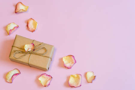 Gift box wrapping in kraft paper with dried cream rose petals on pink pastel background. Love, romance or Valentine's day concept. Flat lay, copy space, top viewの写真素材