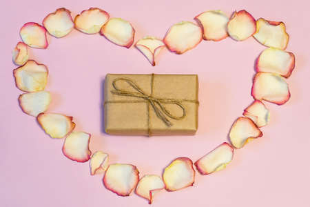 Gift box wrapping in kraft paper in frame of dried cream rose petals in heart shape on pink pastel background. Love, romance or Valentine's day concept, mock up. Flat lay, copy space, top viewの写真素材