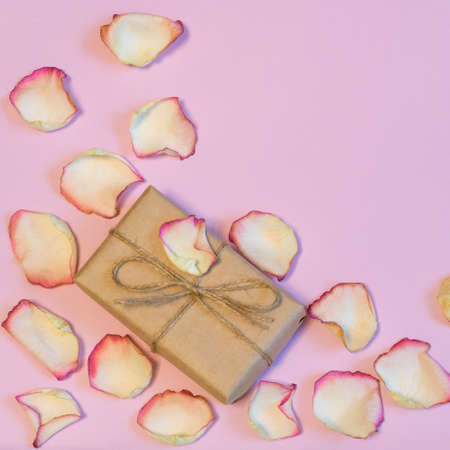Gift box wrapping in kraft paper with dried cream rose petals on pink pastel background. Love, romance or Valentine's day concept. Flat lay, copy space, top viewの写真素材