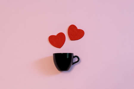 Black cup for coffee or tea and two red hearts on pink pastel background. Creative layout in minimal...