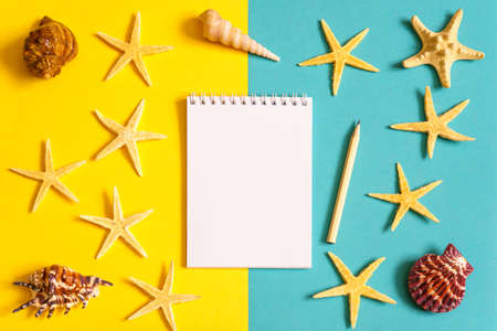 Notebook and pencil with seashells and sea stars on blue and yellow paper background. Mock up, flat lay, copy space, top view. Summer vacation conceptの写真素材
