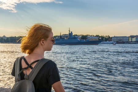 Redhead tween tourist girl on Neva river embankment looking at warships and architectural ensemble in summer evening at sunset. Authentic lifestyle moments, travel concept. Saint Petersburg, Russia.の写真素材