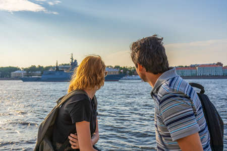 Middle-aged dark-haired man and young redhead lady on Neva river embankment looking at warships and architectural ensemble in summer evening at sunset. Travel concept. Saint Petersburg, Russia.の写真素材
