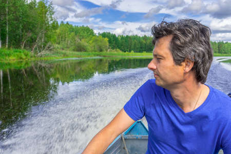 Handsome middle-aged man sitting at boat stern, floating along northern river and relaxing on...
