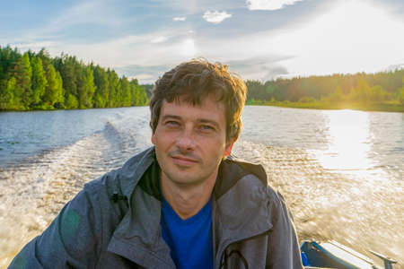 Handsome middle-aged man sitting at boat stern and floating along northern river on beautiful...