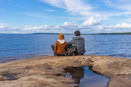 Tourists man and woman sitting on northern lake shore in summer day. People relaxing and admiring beautiful landscape. Travelling and discovering distant places of Earth. Onega lake, Karelia, Russiaの写真素材