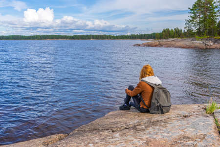 Young tourist lady sitting on northern lake shore in summer day. Teenage girl relaxing and admiring beautiful landscape. Travelling and discovering distant places of Earth. Onega lake, Karelia Russiaの写真素材