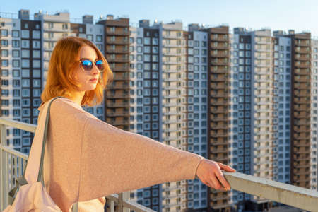 Tween redhead girl in pullover, jeans and sunglasses standing on balcony against high-rise...