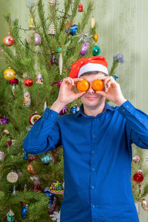 Handsome middle-aged man having fun against beautiful decorated new year tree. Man celebrating Christmas with mandarin oranges. Winter holidays at cozy home concept.の写真素材