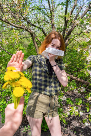 Season allergy to flowering plants pollen. Young woman with paper handkerchief covering her nose in garden and doing stop sign to dandelion bouquet. Teen girl sneezing against blossoming trees.の写真素材
