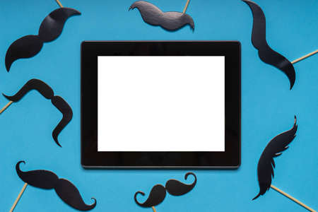 Various black photo booth props moustaches and empty tablet screen on blue background. Greeting card for father's day or men's health awareness month campaign concept. Flat lay, copy space, mock up.の写真素材