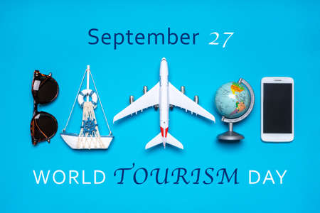 Happy world tourism day. Touristic objects, smart phone, sunglasses, globe and decorative airplane and vessel on blue background. Flat lay, top view. Text September 27, WORLD TOURISM DAY.の写真素材