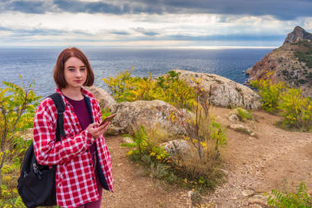 Teen tourist girl with smartphone standing against beautiful seascape in cloudy day. Young lady posing on background of sea, mountains and sky. Traveling and discovering distant places of Earth.の写真素材