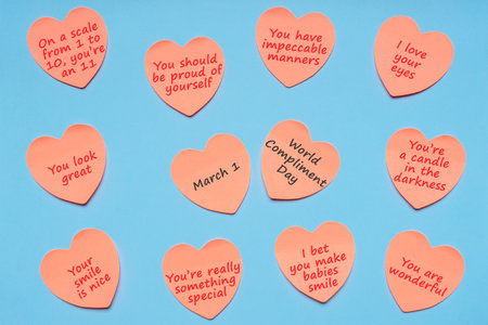 Happy World Compliment Day. Pink paper stickers in heart shape with text of popular compliments for beautiful lady on blue pastel background. Text 1 MARCH WORLD COMPLIMENT DAY. Flat lay, top view.の写真素材