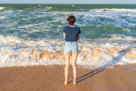 Tween slender girl standing on sand beach of sea coast against stormy waves in sunny summer day....