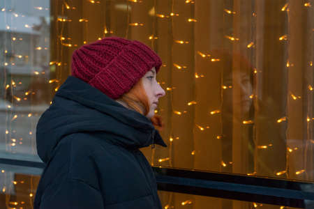 Young beautiful lady near window decorated with Christmas garland at city street in winter. Teen...