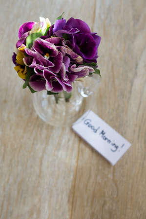 Viola flowers in a vase, old-styled clock and Good morning note on the tableの写真素材