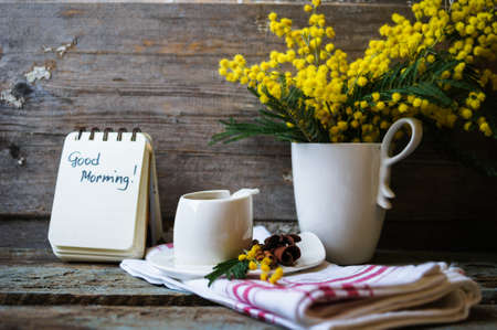 Glass cup with tea and blooming yellow mimosa with good morning note on wooden background