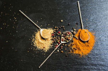 Different types of pepper spice in vintage spoons on black stone background