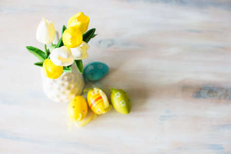 Bright yellow and white tulips in a vase with colorful easter eggs on white rustic wooden table