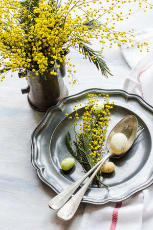 Festive table setting for Easter dinner with bright easter eggs and mimosa flowers on rustic plateの写真素材