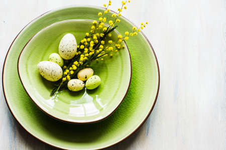 Festive table setting for Easter dinner with bright easter eggs and mimosa flowers on rustic plateの写真素材