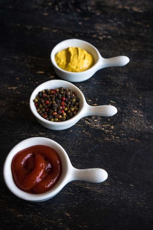 Three bowls with sauces - mustard, ketchup and pepper grain on dark wooden tableの写真素材