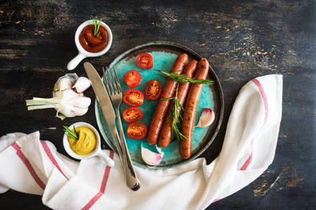 Unhealthy food - grilled sausages with rosemary and tomatoes on dark wooden tableの写真素材
