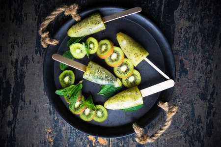 Summer fruit dessert - kiwi lollipops with mint and ice on rustic plate on concrete backgroundの写真素材