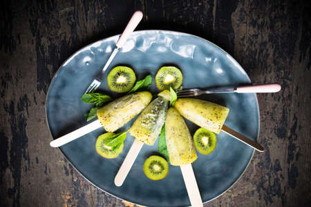 Summer fruit dessert - kiwi lollipops with mint and ice on rustic plate on concrete backgroundの写真素材