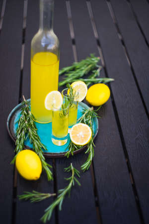 Traditional italian lemon alcohol drink limoncello with pieces of lemon and rosemary herb on dark wooden tableの写真素材