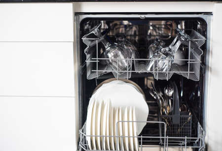 Open dishwasher with clean dishes and wine glasses in the white modern kitchen
