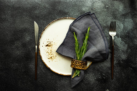 Table setting with ceramic plate and cutlery decorated with fresh rosemary herb on dark stone...