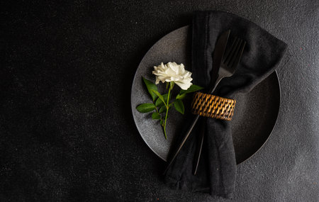 Minimalistic table setting with black stoneware and cutlery decorated with white rose flower on black stone table with copy spaceの写真素材