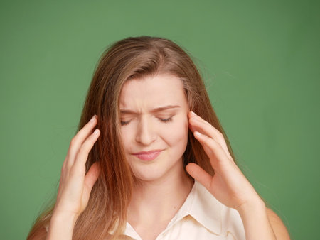 Close-up portrait of a young woman, clutching her head, fingers in hair, eyes lowered, feeling unwell. Isolated on a green background. Frontal full faceの写真素材