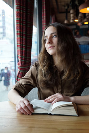 with book and looks in the window, thinking. Dark-brown eyes, mouth closed, natural make-up. Concept: romantic relationship, head in the cloudsの写真素材