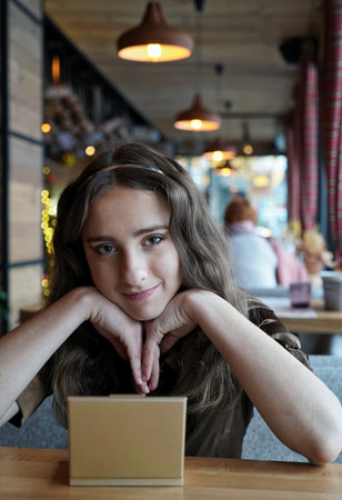 Young long-haired girl with bokeh on the backdrop. Curly haired woman sits in coffee shop and smils. She leans her chin on her handsの写真素材