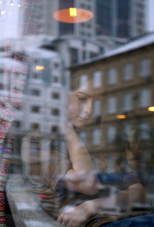 Young girl waiting in cafe for someone, who late. Her hand near chin, head bowed. The window displays the old house and office building. City life, window reflection, urban styleの写真素材