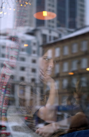 Young european woman waiting for someone, who late. Her hand near chin. The window displays the old house and office building. City life, window reflection, urban styleの写真素材