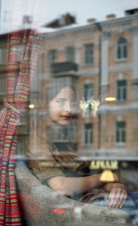 A frustrated girl sits behind the window of cafe. She is concerned about something, her hands are...