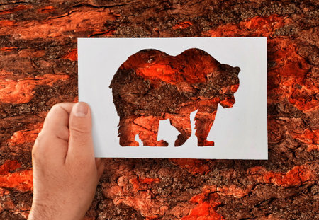 Man, flora, fauna. A hand holds a paper stencil of bear against background of tree bark. The...