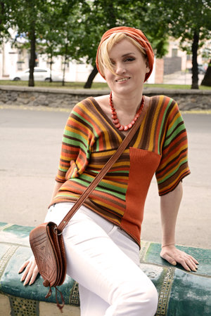 Style of the 1970s. A beautiful long-haired blonde in retro clothes is sitting on a bench in the city: white tights, model hair, high heels, motley accessories, striped blouseの写真素材