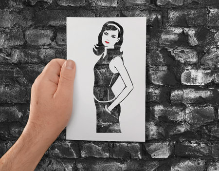 A hand holds a paper stencil of young long-haired girl against background of black bricks. The contours of beautiful woman. Fashion, glamour. Dress making, dress patternの写真素材