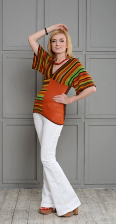 Beautiful girl sitting on a black cube in 1970s clothes. She is dressed in a striped knitted blouse and crocheted white jeans, high heels, hand up and on the waist.の写真素材