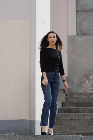 A beautiful woman in jeans and a black blouse - T-shirt in city on the street near wall. The glasses in hand, high heals. Dark hair develops in the wind. Street fashion, denim style, summer - autumnの写真素材